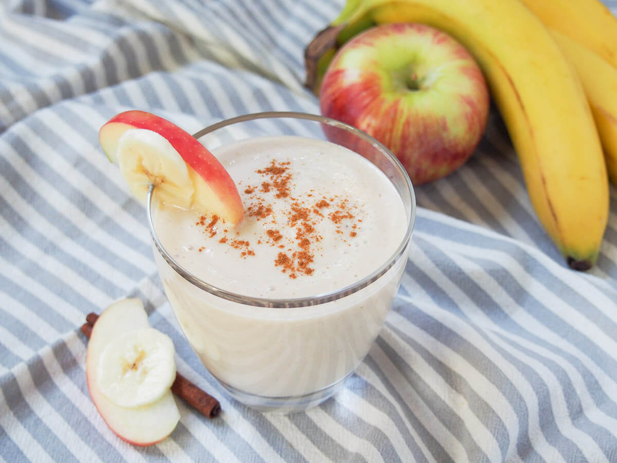 glass of apple banana smoothie with apple and bananas in background behind/to side.