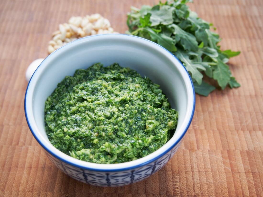 bowl of arugula pesto with arugula and pine nuts behind