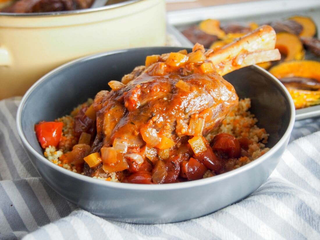 braised lamb shank in bowl with sauce over top and pot it was cooked in behind