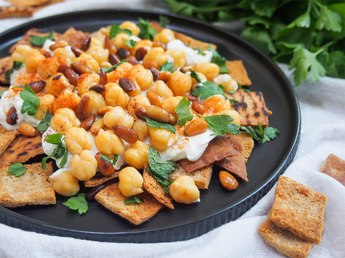 plate of chickpea fatteh with extra toasted pita to side and herbs behind