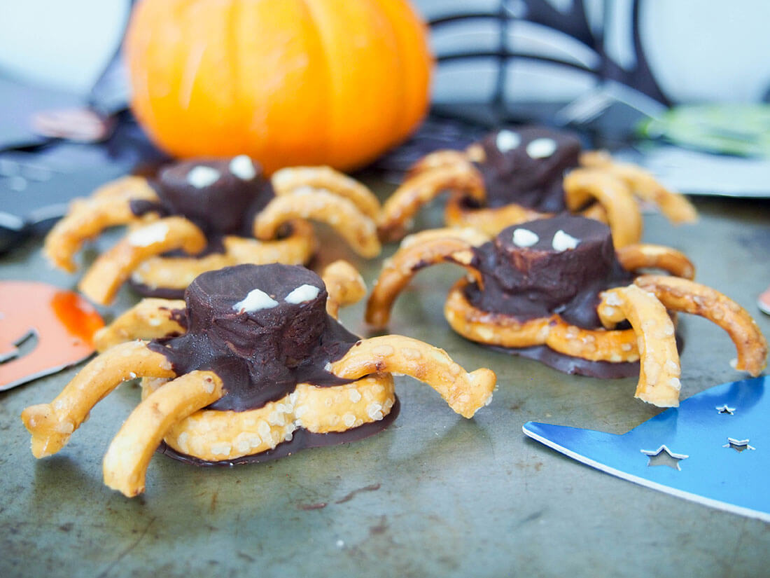 four chocolate pretzel spiders with pumpkin and spider web cutout blurred behind