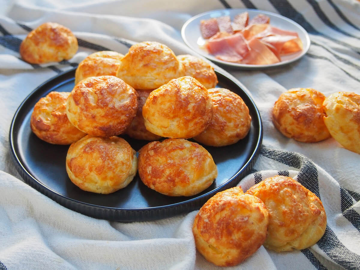 plate of gougeres cheese puffs with some in front and small plate of ham behind