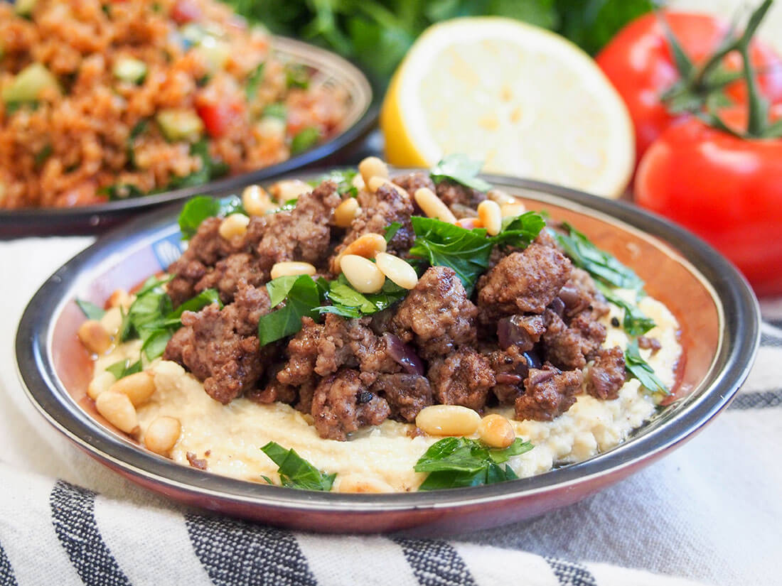 Hummus kawarma (hummus with lamb) in small dish with tomatoes and plate of kisir behind