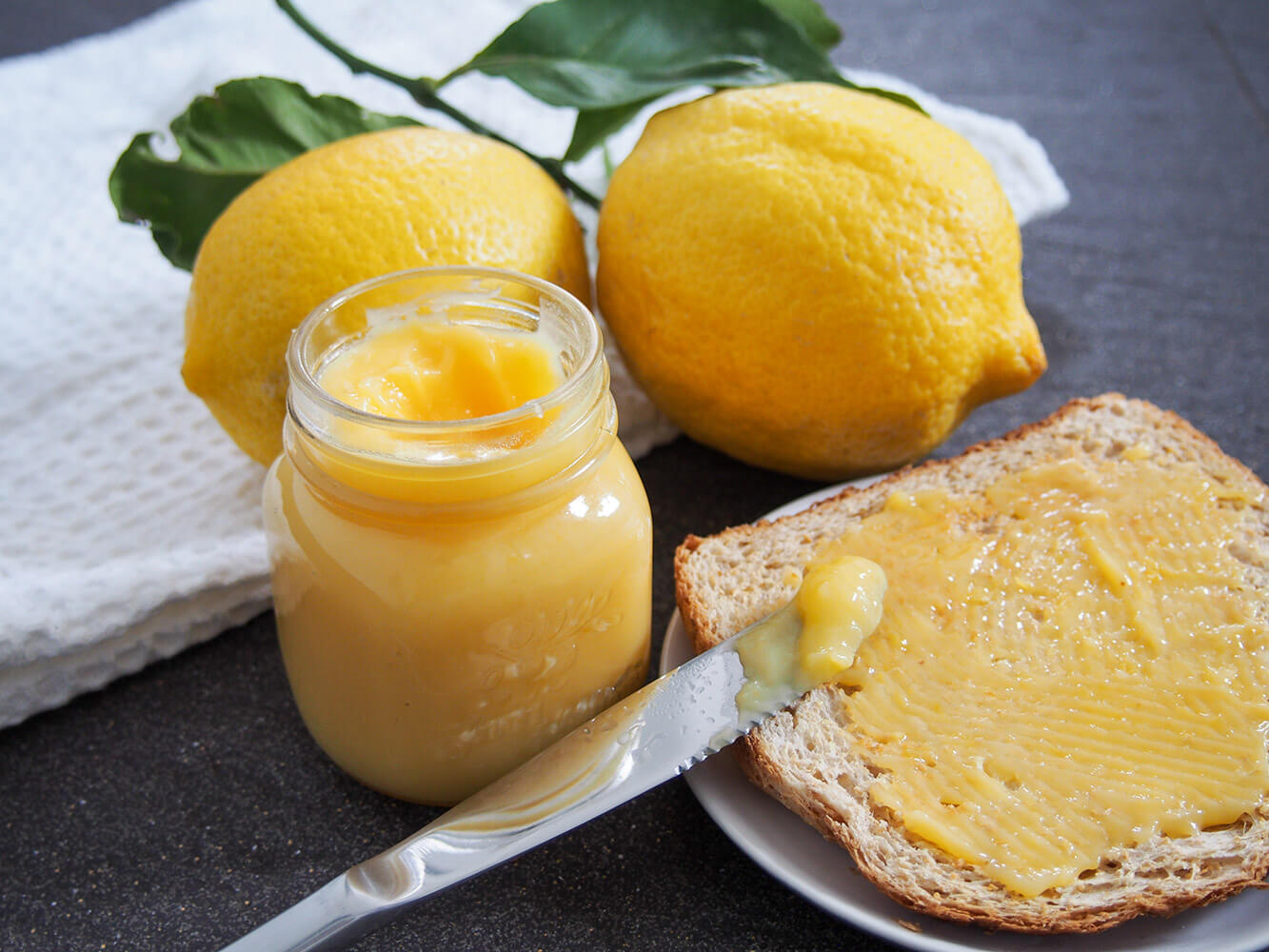 jar of lemon curd with some on toast to side and lemons behind