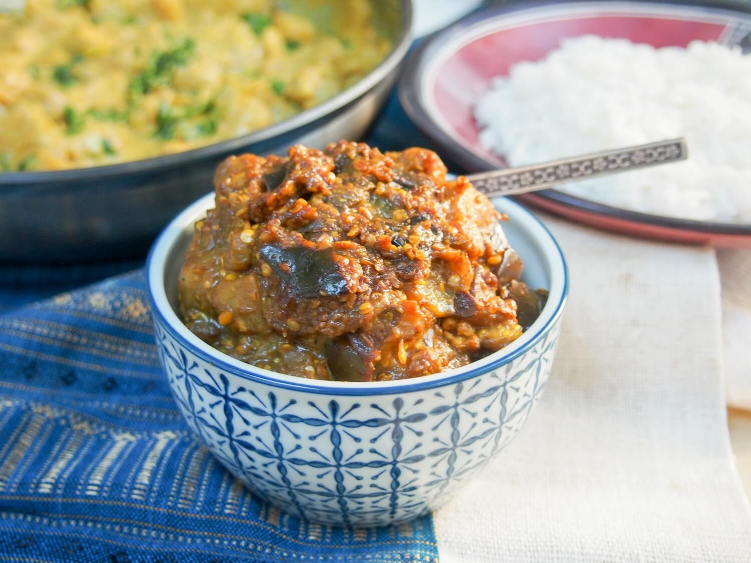 brinjal pickle (Indian eggplant relish/aubergine chutney) with spoon in dish and plates of curry and rice behind