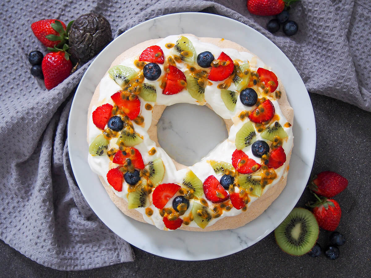 pavlova wreath topped with cream and fruit with additional fruit to side of plate