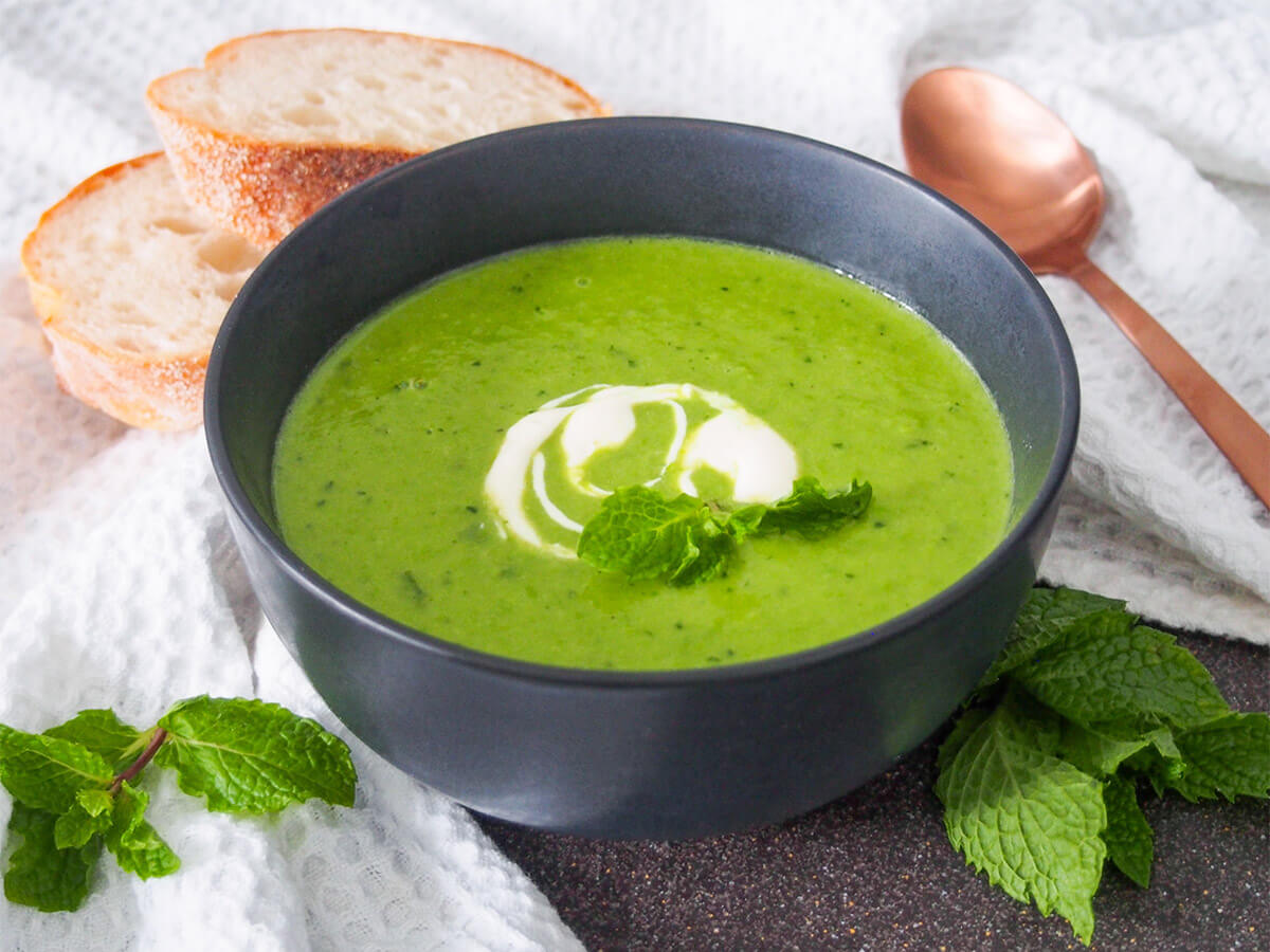 bowl of pea and mint soup with spoon and bread behind and mint sprigs to side of bowl