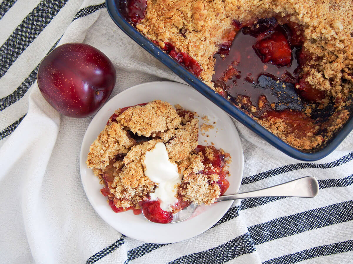 plate with plum crumble topped with a little cream with a spoon on bottom of plate, rest of crumble to side and plum to other side of plate