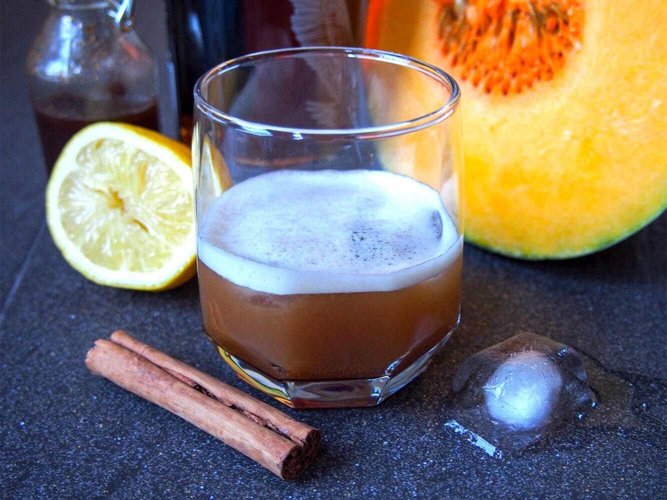 glass of pumpkin spice whiskey sour with cinnamon stick, lemon and pumpkin around glass