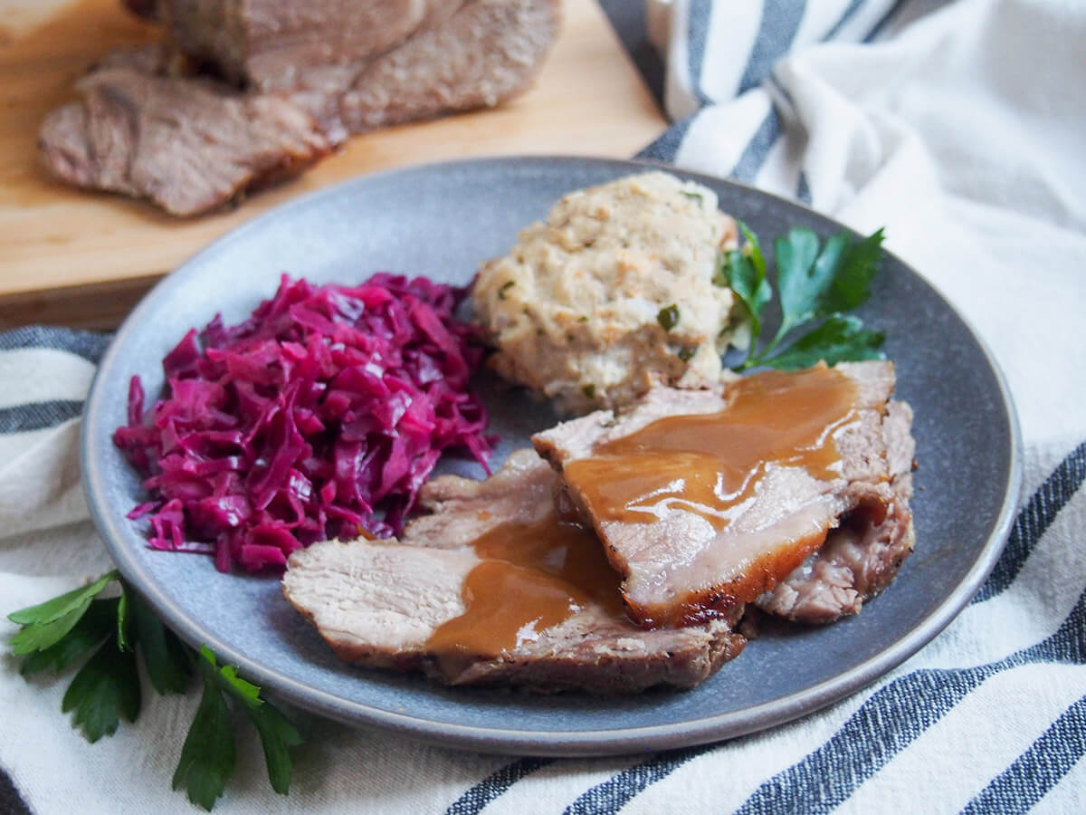 slices of Schweinebraten - German pork roast - on plate with red cabbage and dumpling behind