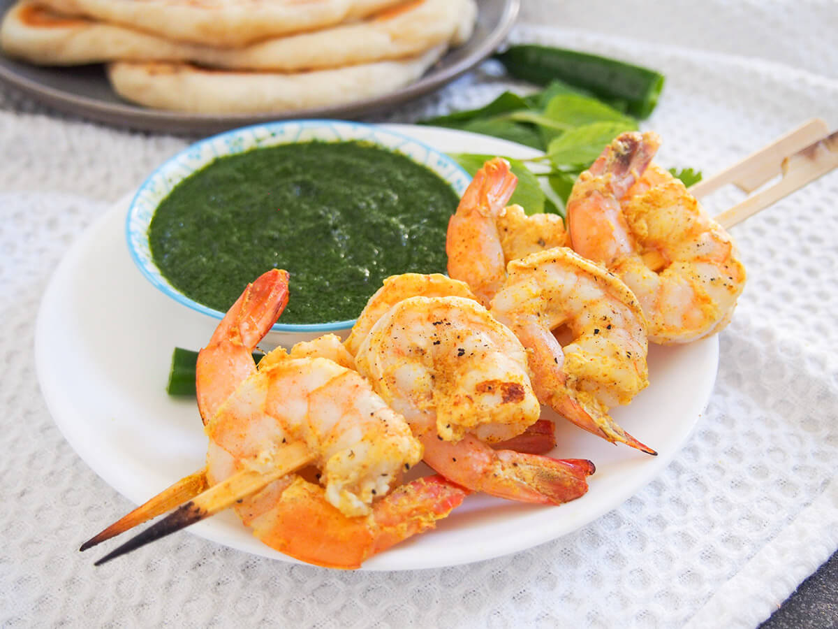 two skewers of tandoori shrimp with mint chutney behind and naan in background