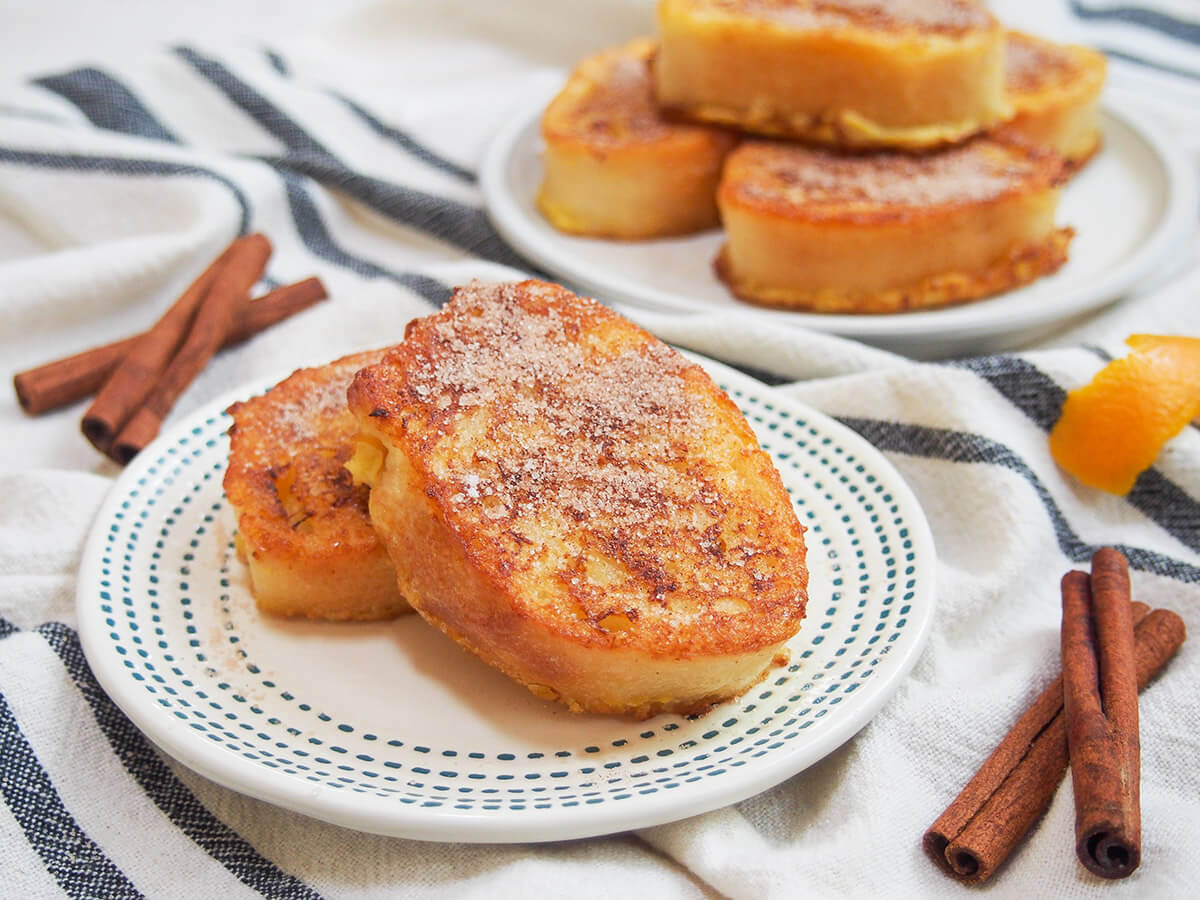 slices of torrijas on plate with cinnamon sticks to side and plate with stack more behind